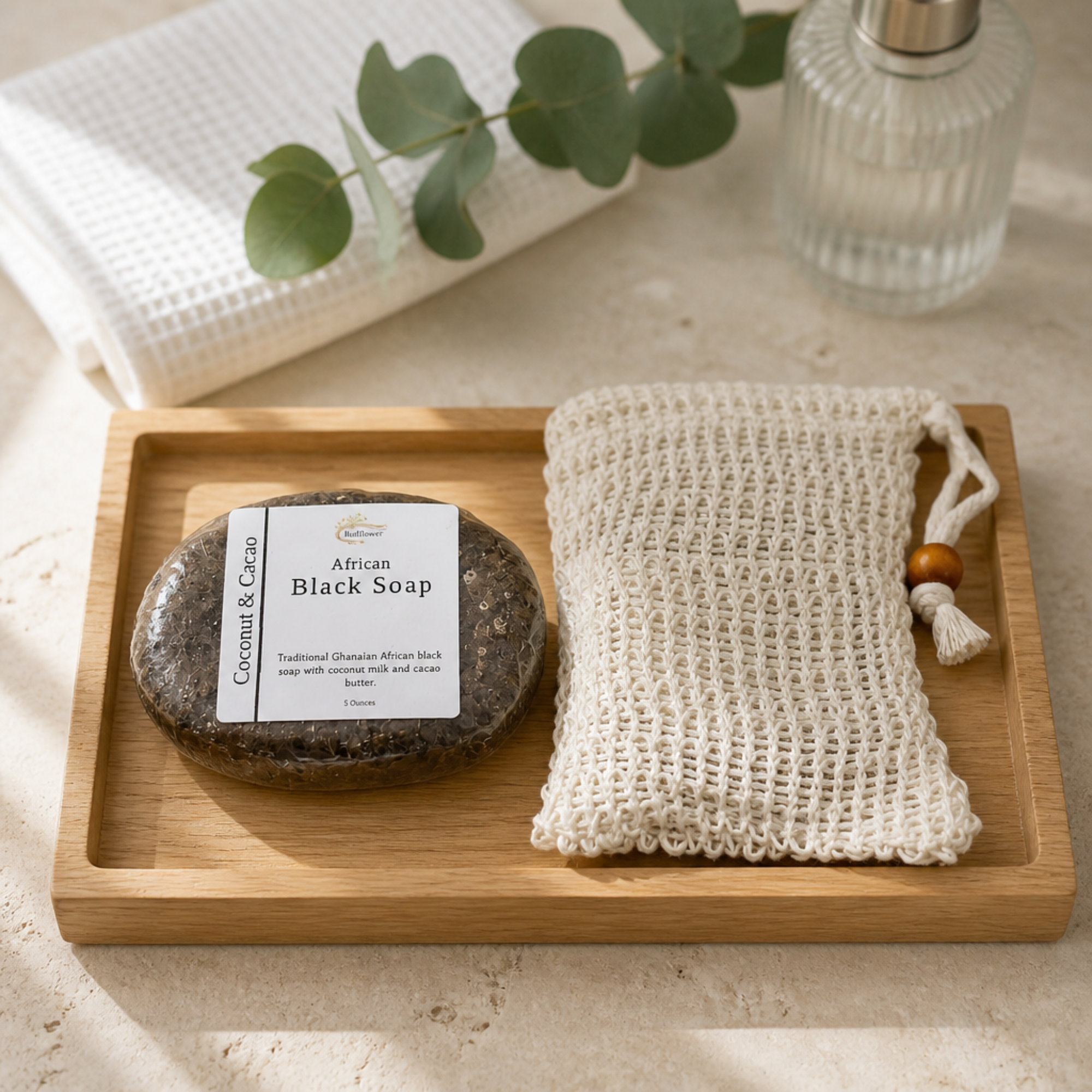 African Black Soap and a loofah on a wooden tray with a towel and bottle in the background.